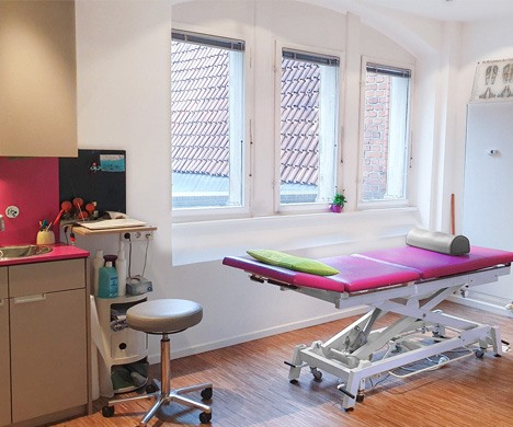 Behandlungsraum einer Physiotherapiepraxis mit verstellbarer Liege, Hocker und großen Fenstern